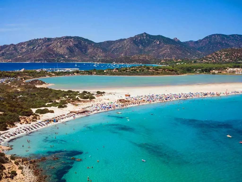 Acque turchesi cristalline baciano una spiaggia assolata piena di vacanzieri al VOI Tanka Resort, mentre uno sfondo di aspre montagne completa questo idilliaco paradiso costiero in Sardegna.