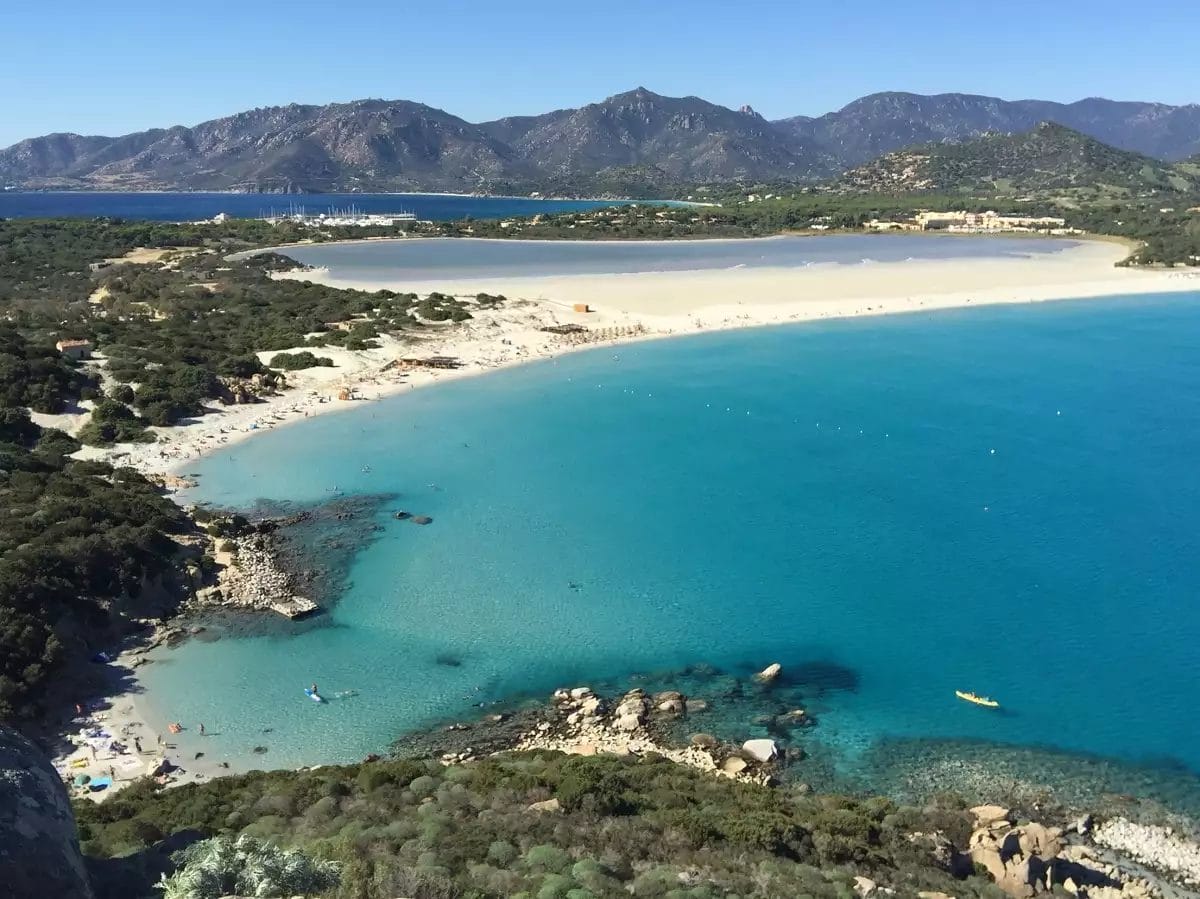 Una vista pittoresca di una serena baia turchese in Sardegna, con una spiaggia di sabbia bianca bordata da una vegetazione lussureggiante e rocce aspre, sotto un cielo azzurro e limpido vicino al VOI Tanka Resort.