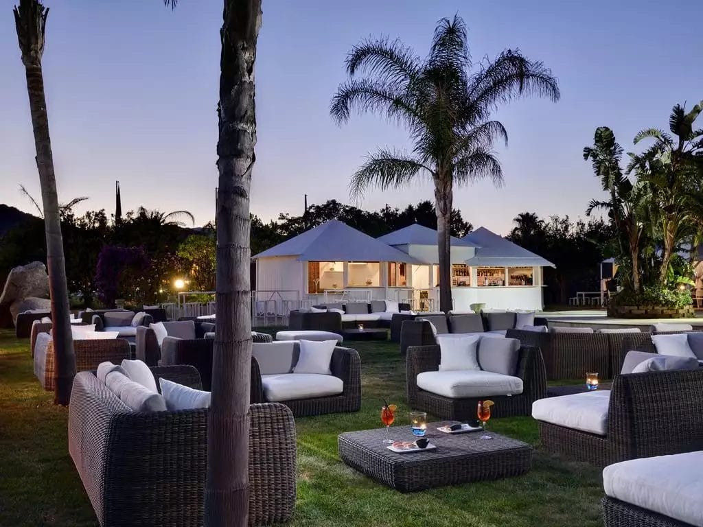 Serenità crepuscolare in una lussuosa lounge all'aperto del VOI Tanka Resort in Sardegna, con lussuosi posti a sedere, palme tropicali e un invitante padiglione bar sotto un limpido cielo serale.