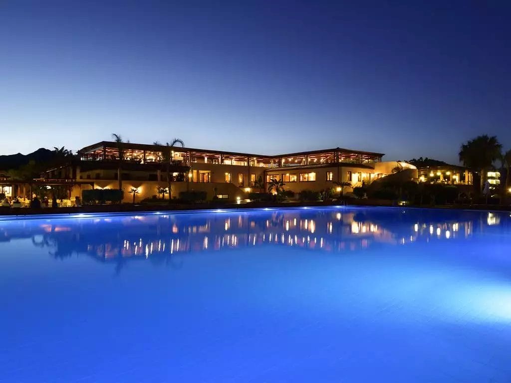 Serata tranquilla al VOI Tanka Resort con una serena piscina illuminata di blu che riflette il caldo bagliore della facciata di un grand hotel sotto il cielo al crepuscolo.