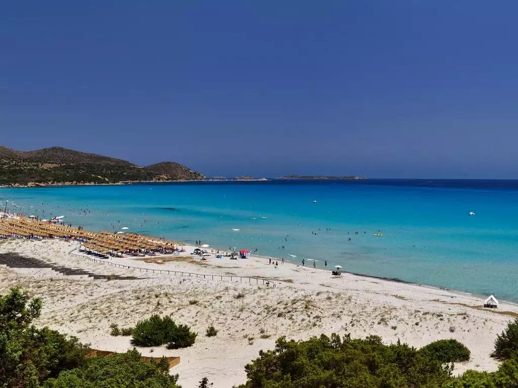 Una spiaggia serena al VOI Tanka Resort con acque turchesi cristalline, una spiaggia sabbiosa punteggiata da ombrelloni colorati e bagnanti, con uno sfondo di colline sotto un cielo azzurro e limpido.