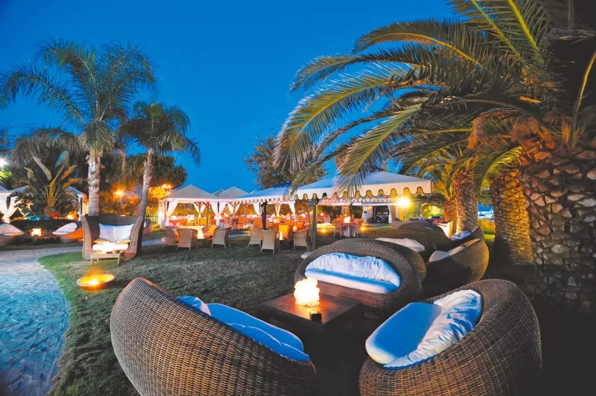 Una serata in un tranquillo resort di lusso in Sardegna, con un'atmosfera calda e invitante, mentre gli ospiti cenano sotto una tenda circondata da palme e accoglienti aree lounge con lussuosi posti a sedere.