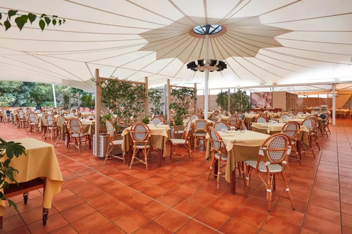 Una zona pranzo all'aperto aperta e ariosa al VOI Tanka Resort con grandi ombrelloni che offrono ombra, con tavoli elegantemente apparecchiati e sedie in vimini su un pavimento in piastrelle di terracotta.