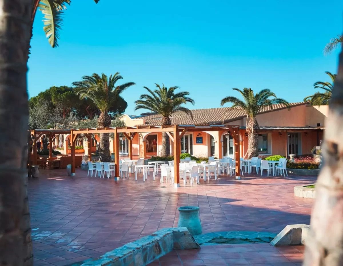 Zona pranzo all'aperto nel patio con tavoli e sedie bianchi sullo sfondo di edifici dal tetto in terracotta e rigogliose palme sotto un cielo azzurro e limpido presso il lussuoso VOI Tanka Resort a S.