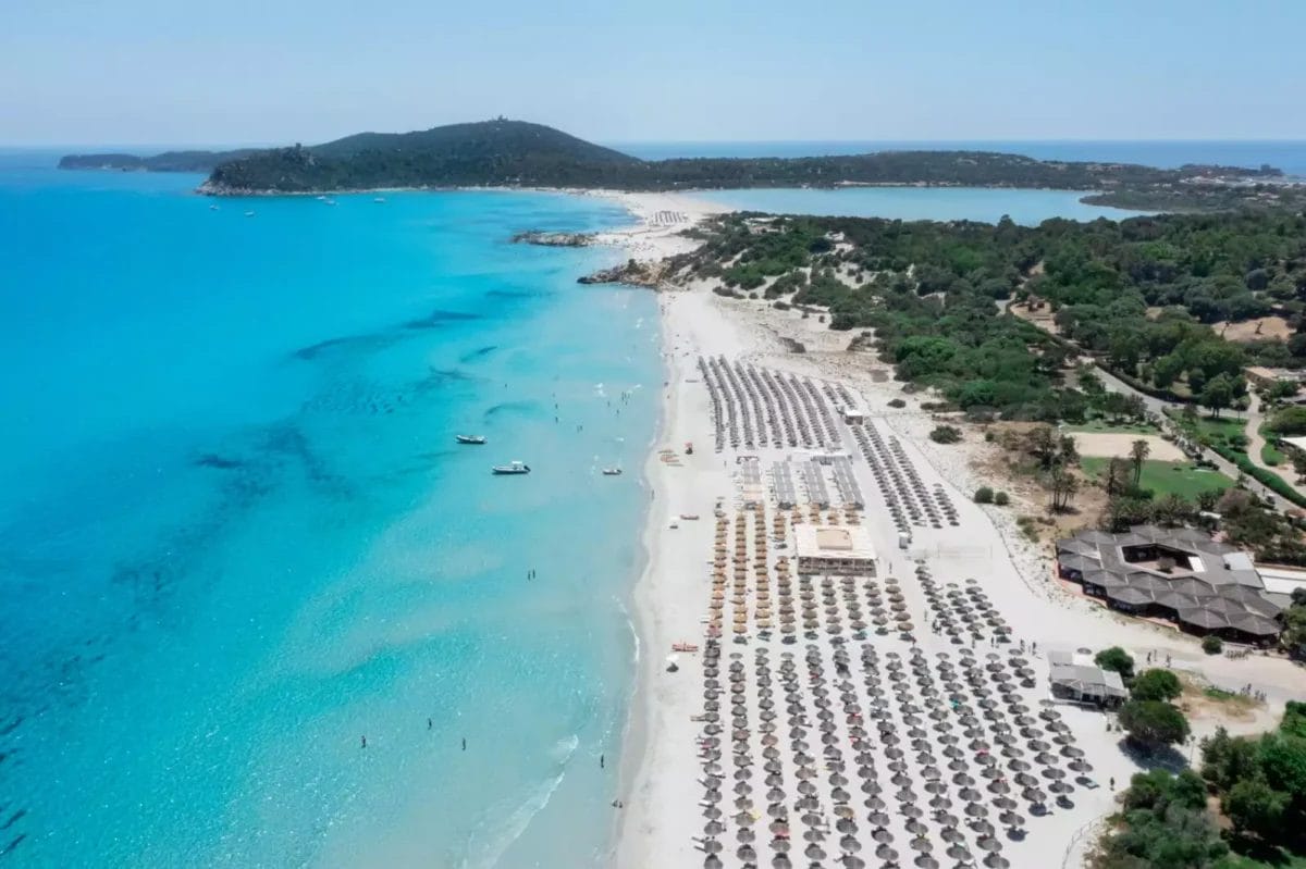 Una veduta aerea del VOI Tanka Resort mostra una spiaggia incontaminata con acque turchesi cristalline, fiancheggiata da file di lettini e ombrelloni, sullo sfondo di una vegetazione lussureggiante a S