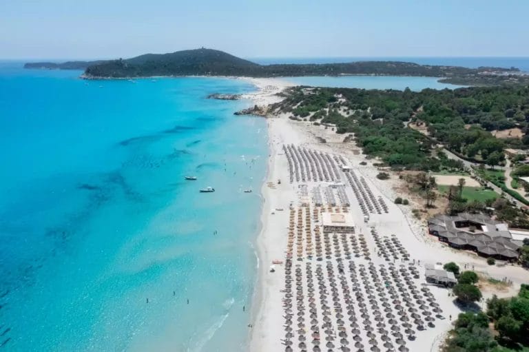 Veduta aerea di una tranquilla spiaggia tropicale in Sardegna con acque turchesi cristalline, una distesa di sabbia bianca fiancheggiata da file di lettini e ombrelloni nel resort di lusso, VOI