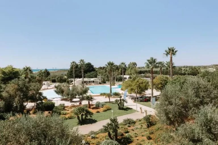 Una vista serena del Club La Giurlita, un lussureggiante resort con piscine circondate da palme e fitta vegetazione, affacciato sul mare.