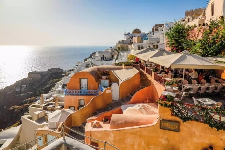 Vista al tramonto di Oia, Santorini, con edifici bianchi e tetti in terracotta che si affacciano sul mare, con caffè all'aperto e un mulino a vento in lontananza. Questa destinazione di viaggio offre viste