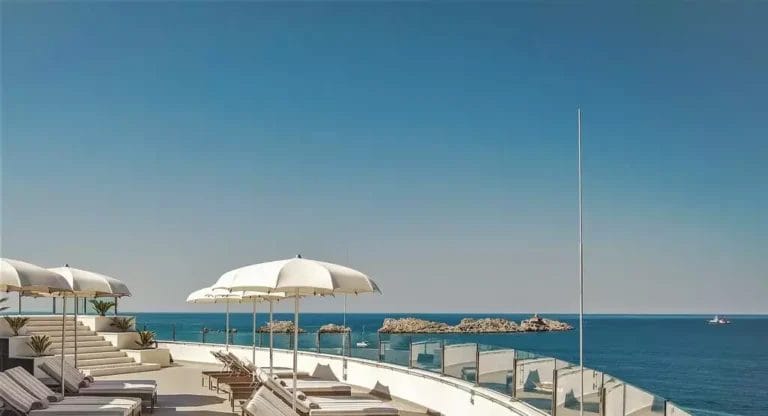 Un'area lounge sul mare con lettini e ombrelloni bianchi che si affaccia su un oceano cristallino con affioramento roccioso visibile, fornita dal Royal Blue Hotel.