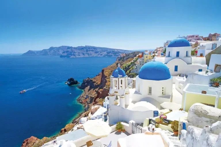 Veduta di Santorini con le iconiche chiese dalla cupola blu, edifici bianchi e il Mar Egeo sotto un cielo azzurro con Alpiselect Afroditi.