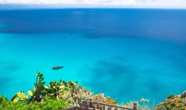 Una vista panoramica di un mare turchese e limpido da una scogliera costiera, con i cactus Opuntia in primo piano e una piccola barca nell'acqua, simbolo del viaggio ecosostenibile.