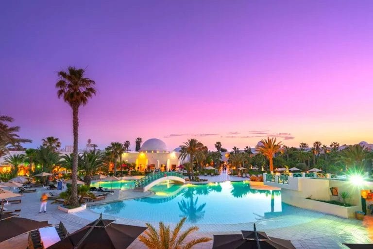 La piscina di un hotel con cupola bianca e palme durante il tramonto, caratterizzata da vivaci cieli viola e ambienti illuminati.