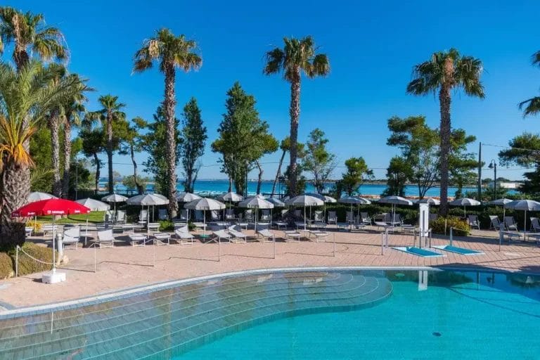 Una zona piscina con lettini, ombrelloni e una vista sulla spiaggia del Salento adornata da palme e pini sotto un cielo azzurro e limpido, offre un fascino sereno da villaggio.