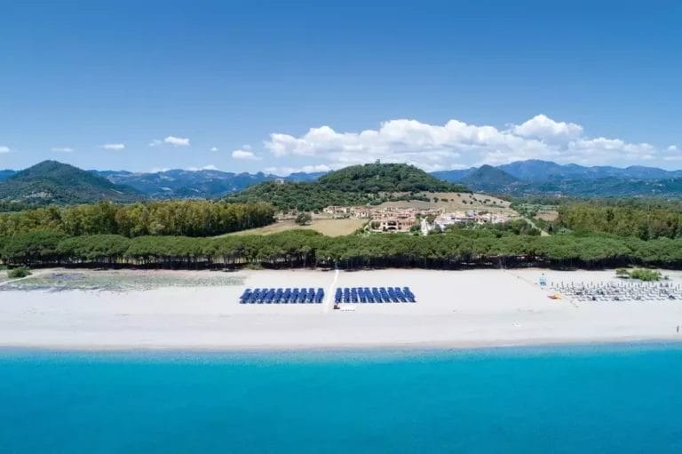 Veduta aerea del lungomare del Cala Luas Resort con file di ombrelloni blu e sedie a sdraio. Dietro la spiaggia ci sono alberi, edifici e terreno collinare sotto un cielo azzurro e limpido.