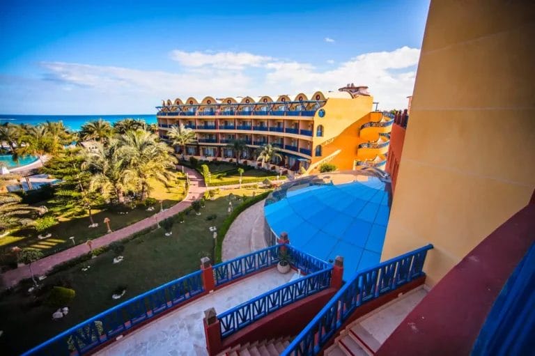 Vista di Beau Rivage, una colorata località balneare con un edificio a più piani, giardini lussureggianti, una scalinata e il mare sullo sfondo in una giornata limpida e soleggiata.