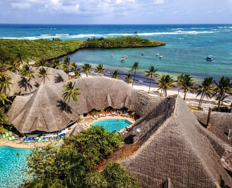 Veduta aerea dell'IGV Club, un resort tropicale con edifici dal tetto di paglia, due piscine e palme lungo una splendida costa. Le barche ancorano vicino a un'isola al largo circondata dalle acque scintillanti di Blue Bay.