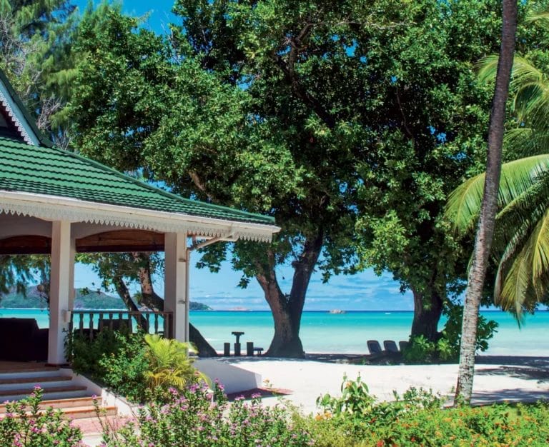 Un gazebo fronte mare a Cote d'Or si trova in mezzo a una vegetazione lussureggiante, affacciato su una spiaggia sabbiosa con acque cristalline e isole lontane sotto un cielo luminoso e soleggiato.