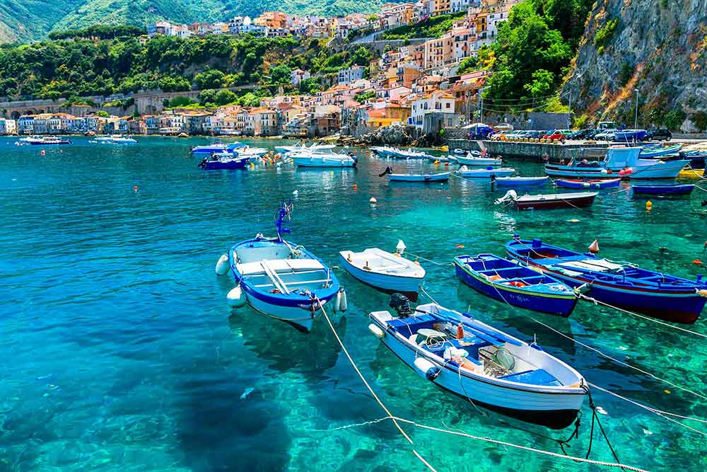 Piccole imbarcazioni ancorate in un porto azzurro con case colorate e verdi colline sullo sfondo, che lo rendono una pittoresca destinazione di viaggio in Italia.