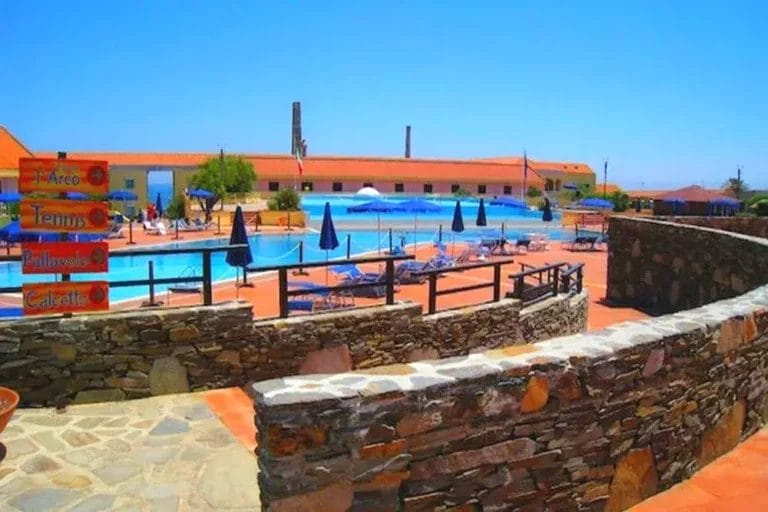 Area piscina all'aperto con sedie a sdraio e ombrelloni, circondata da un sentiero in pietra e un muro. La segnaletica indica vari servizi tra cui tiro con l'arco, tennis e una caffetteria. Cielo azzurro e limpido in alto. Vivi il lusso al resort sulla spiaggia Le Tonnare a Stintino.