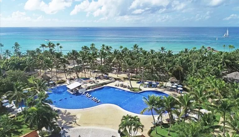 Un'ampia area piscina all'aperto con sedie a sdraio circondate da palme. Oltre la piscina del Catalonia Gran Dominicus, una spiaggia sabbiosa conduce all'oceano con barche a vela all'orizzonte sotto un cielo parzialmente nuvoloso.