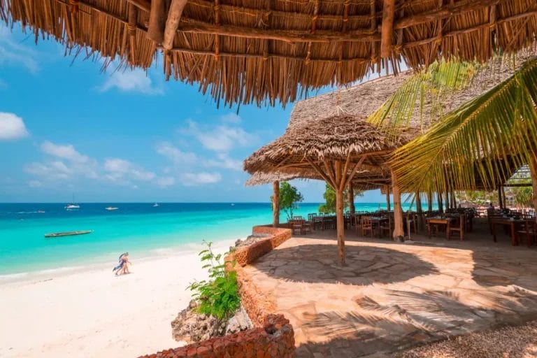 Resort sulla spiaggia con strutture con tetto di paglia e tavoli e sedie in legno su una spiaggia sabbiosa. Una coppia cammina lungo la riva, mentre le barche galleggiano sull'acqua turchese sotto un cielo parzialmente nuvoloso al Sandies Seaclub.