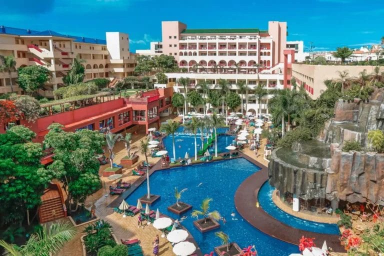 Vista aerea del resort Alpiclub con una grande area piscina con sedie a sdraio e ombrelloni, circondata da vegetazione tropicale e edifici a più piani sullo sfondo sotto un cielo azzurro terso. I colori vivaci degli alberi di jacaranda aggiungono un tocco di paradiso in più a questa fuga idilliaca.