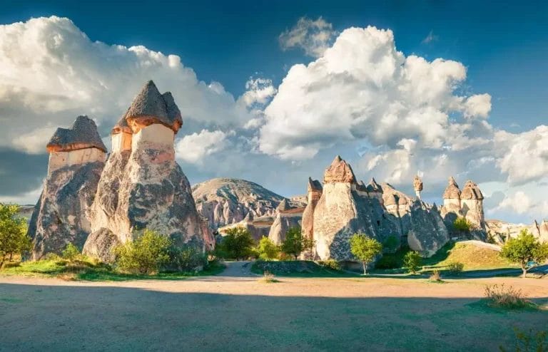 Vista panoramica di formazioni rocciose uniche sotto un cielo parzialmente nuvoloso in Cappadocia, Turchia, con alberi e sentieri in primo piano: una tappa obbligata del tuo GRAN TOUR DELLA TURCHIA.