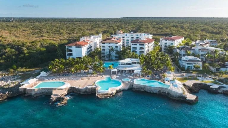 Vista aerea dell'HM Alma de Bayahibe Resort, un paradiso costiero con numerose piscine, edifici bianchi e una vegetazione lussureggiante sullo sfondo di una foresta che si estende fino all'orizzonte; in primo piano l'acqua azzurra dell'oceano.