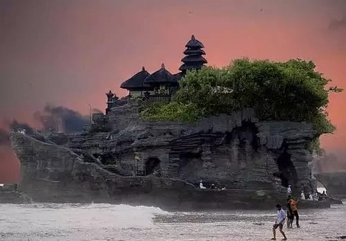 Un tempio su un'isola rocciosa a Nusa Dua, con persone in primo piano, sullo sfondo di un suggestivo cielo arancione.