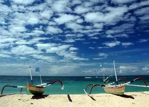 Due barche da pesca tradizionali sono ormeggiate sulla riva sabbiosa di Nusa Dua, sotto un cielo parzialmente nuvoloso e con le acque turchesi dell'oceano sullo sfondo, non lontano da un lussuoso hotel sulla spiaggia e spa.