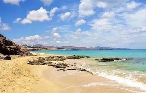 Una spiaggia sabbiosa a Costa Calma con rocce lungo la riva, acqua turchese e limpida e un cielo azzurro parzialmente nuvoloso. Sullo sfondo si vedono colline lontane, che creano un idilliaco scenario costiero.