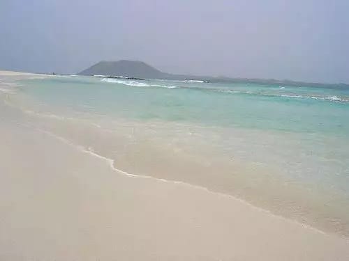 Spiaggia sabbiosa con acque limpide e turchesi e onde dolci; SBH Costa Calma Beach offre viste mozzafiato su un'isola all'orizzonte sotto un cielo nebbioso.