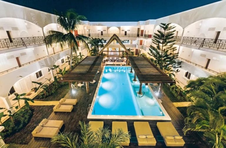 Vista aerea della piscina rettangolare circondata da sedie a sdraio e palme nel cortile di un hotel di notte. I vari piani dell'edificio sono dotati di balconi illuminati, creando un'atmosfera serena perfetta per gli amanti dei viaggi che esplorano Playa del Carmen.