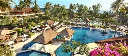 Vista aerea di un lussuoso resort tropicale con numerose piscine, circondato da sedie a sdraio, palme ed edifici, a pochi passi dalla sabbia incontaminata della spiaggia di Nusa Dua.