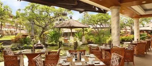 Ristorante all'aperto con tavoli e sedie in legno, con vista su un rigoglioso giardino con laghetto e gazebo sotto un cielo limpido, immerso nella tranquilla atmosfera di un hotel sulla spiaggia di Nusa Dua.