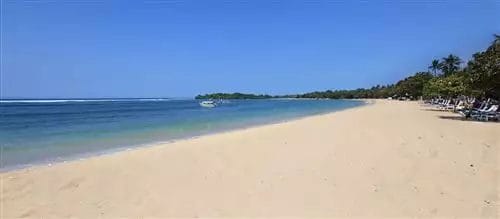 Un'ampia spiaggia sabbiosa si estende lungo le calme e azzurre acque dell'oceano sotto un cielo limpido a Nusa Dua, con una barca in lontananza e alberi che costeggiano la riva vicino a un lussuoso hotel sulla spiaggia.