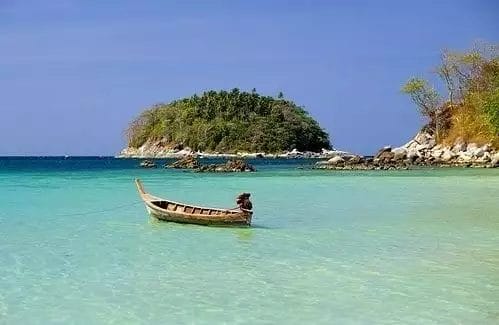 Una piccola barca di legno galleggia sulle limpide acque turchesi vicino a un'isola ricoperta di alberi, sotto il cielo azzurro, catturando la serena bellezza di Capo Panwa come se fosse una cartolina del SeaClub.