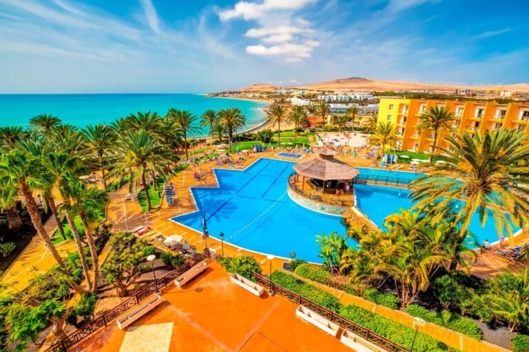 Vista aerea di un resort tropicale a Costa Calma, caratterizzato da una grande piscina blu, palme ed edifici colorati, il tutto con vista sull'oceano turchese sotto un cielo limpido.