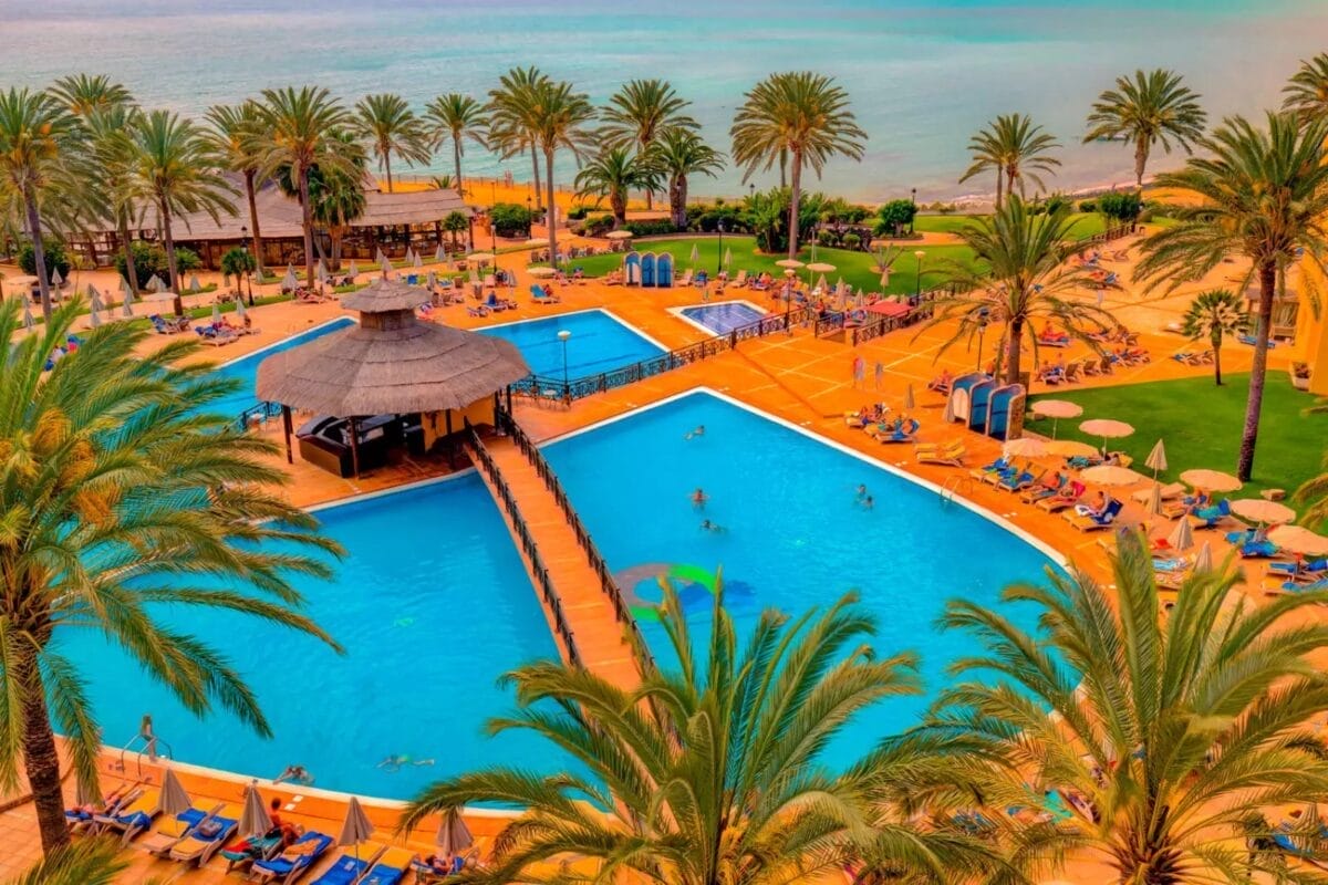 Vista aerea del resort SBH Costa Calma Beach, con due piscine collegate tra rigogliose palme e ospiti che prendono il sole, il tutto incastonato vicino a una spiaggia sabbiosa e all'oceano turchese.