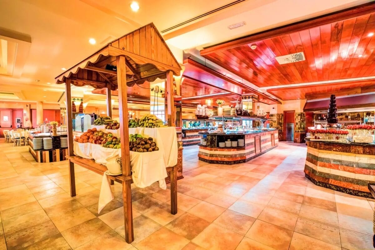 L'area buffet dell'SBH Costa Calma Beach è spaziosa e presenta un carretto di frutta in legno, varie postazioni di ristorazione e un arredamento in legno con luci calde.