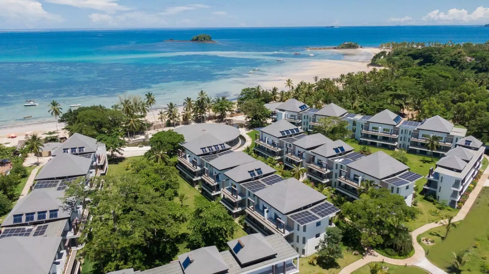 Vista aerea del Royal Andilana Resort, un lussuoso rifugio sulla spiaggia con più edifici circondati da palme e prati verdi lussureggianti, con viste mozzafiato su un oceano blu. Il resort include un SeaClub e una spa indulgente per il massimo relax.