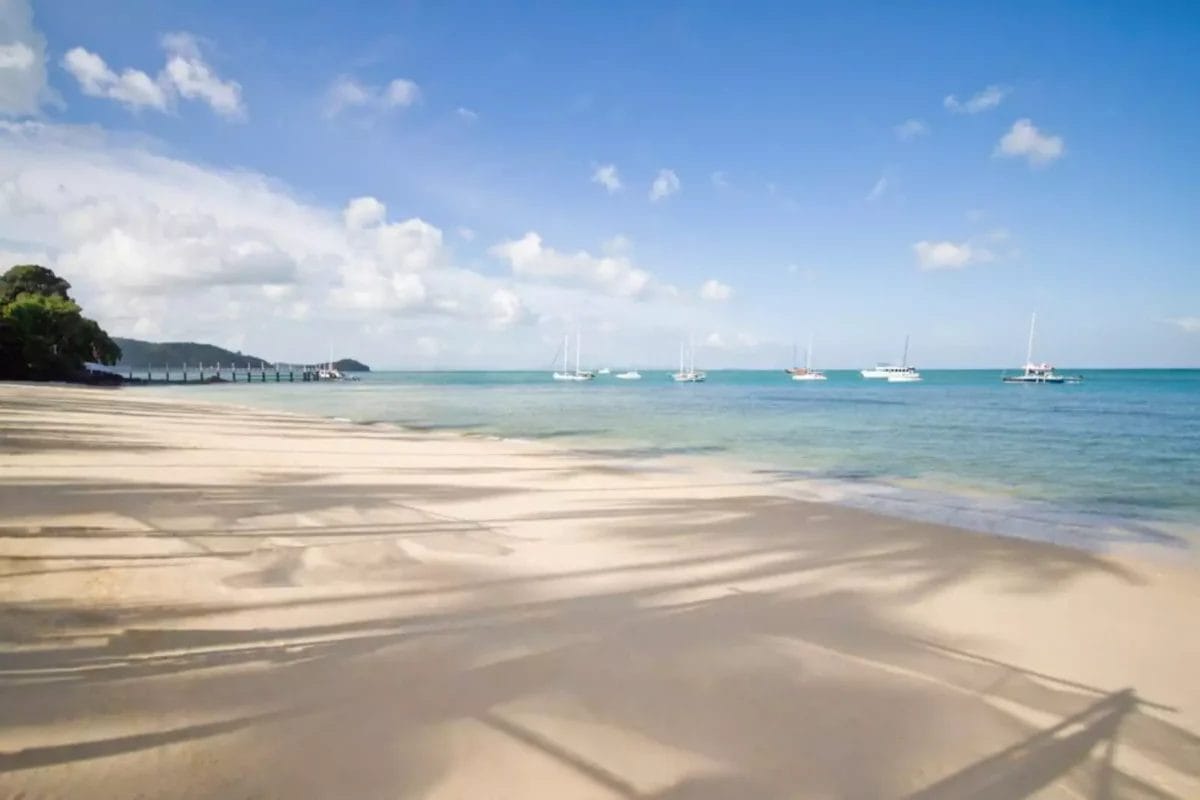 Sotto il cielo terso di Cape Panwa, una spiaggia assolata invita con il suo mare calmo e turchese e le barche a vela ancorate dolcemente in lontananza. Le ombre degli alberi danzano sulla riva sabbiosa, creando una scena pittoresca che ricorda uno stile di mare lussuoso.