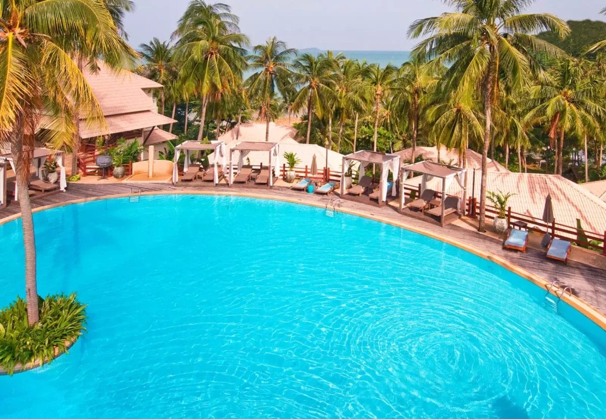 Una grande piscina circolare circondata da lettini e palme vanta una vegetazione tropicale e uno sfondo oceanico in lontananza, richiamando lo stile tranquillo del rinomato SeaClub di Cape Panwa.