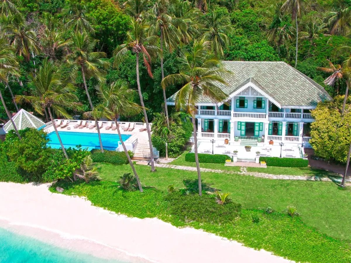 Una vista aerea di un'elegante villa sulla spiaggia di Cape Panwa, con una piscina circondata da palme che conduce a una spiaggia sabbiosa e al mare turchese, evoca il fascino di un esclusivo rifugio in un seaclub.