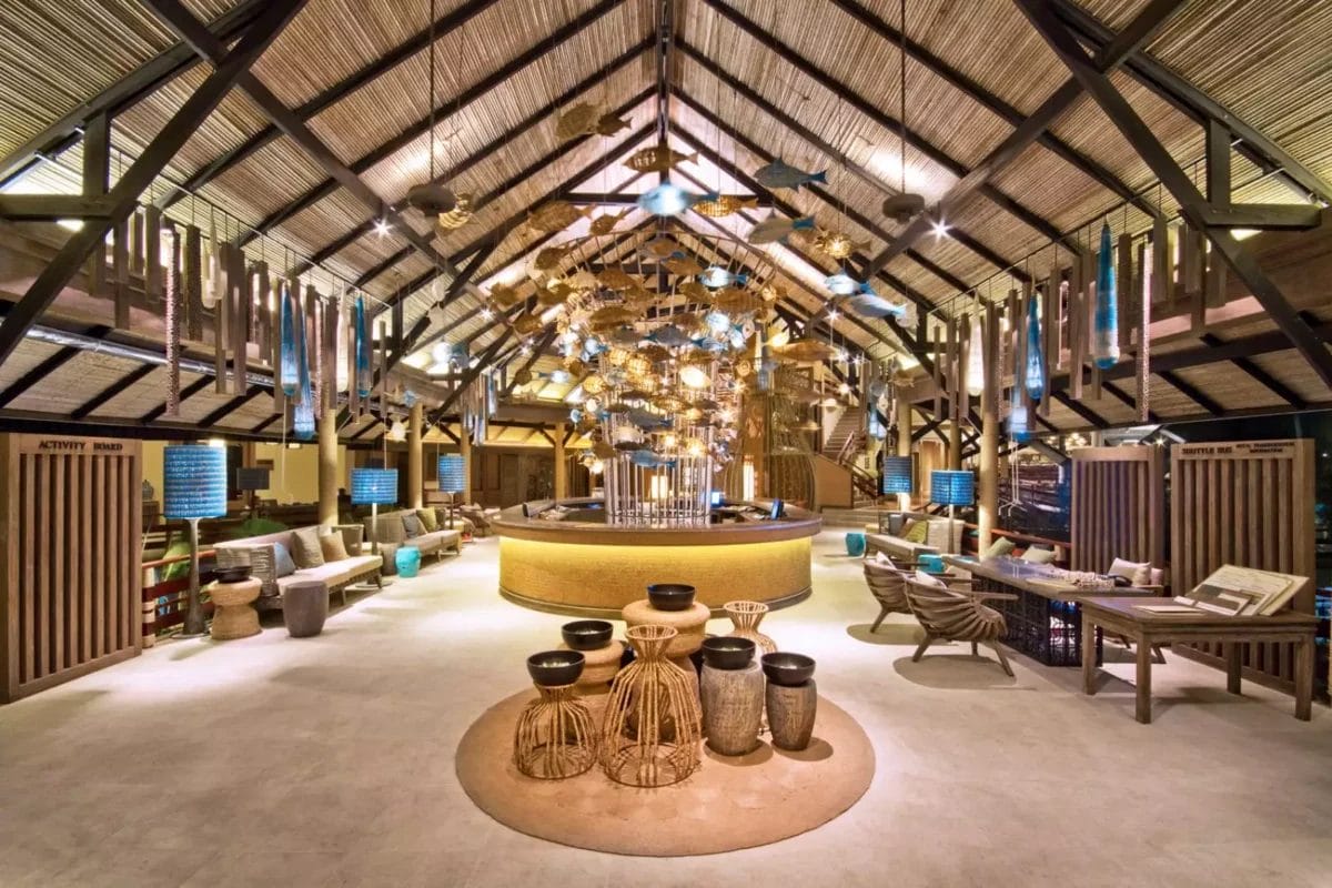 La spaziosa e ben illuminata hall del resort Cape Panwa vanta un alto soffitto in legno e un arredamento moderno con luci sospese. I suoi toni caldi e il bar centrale creano un'atmosfera invitante, riflettendo uno stile unico che promette comfort e relax in ogni area salotto.