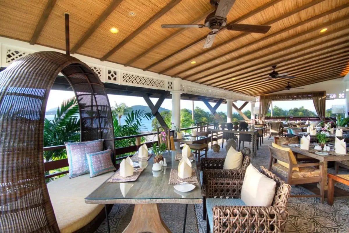 Questo elegante ristorante all'aperto al SEA Club di Cape Panwa è arredato con mobili in vimini, tavoli quadrati apparecchiati per mangiare e una comoda sedia sospesa. Un soffitto in legno esalta l'atmosfera, mentre una vegetazione lussureggiante e colline lontane completano lo sfondo pittoresco.