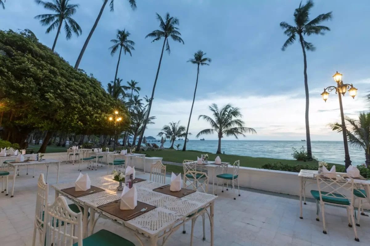 Godetevi un'esperienza culinaria all'aperto in stile sulla spiaggia presso il SEACLUB a Cape Panwa, con tavoli e sedie bianchi sistemati sotto alte palme, di fronte all'oceano al tramonto. Tovaglioli e posate sono elegantemente disposti sui tavoli.