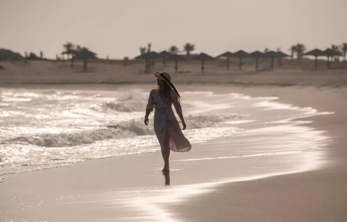 Una persona con un abito e un cappello cammina lungo una spiaggia sabbiosa vicino alla riva sotto un cielo nuvoloso, godendosi l'atmosfera serena che ricorda un'esperienza Oman Esclusivo.