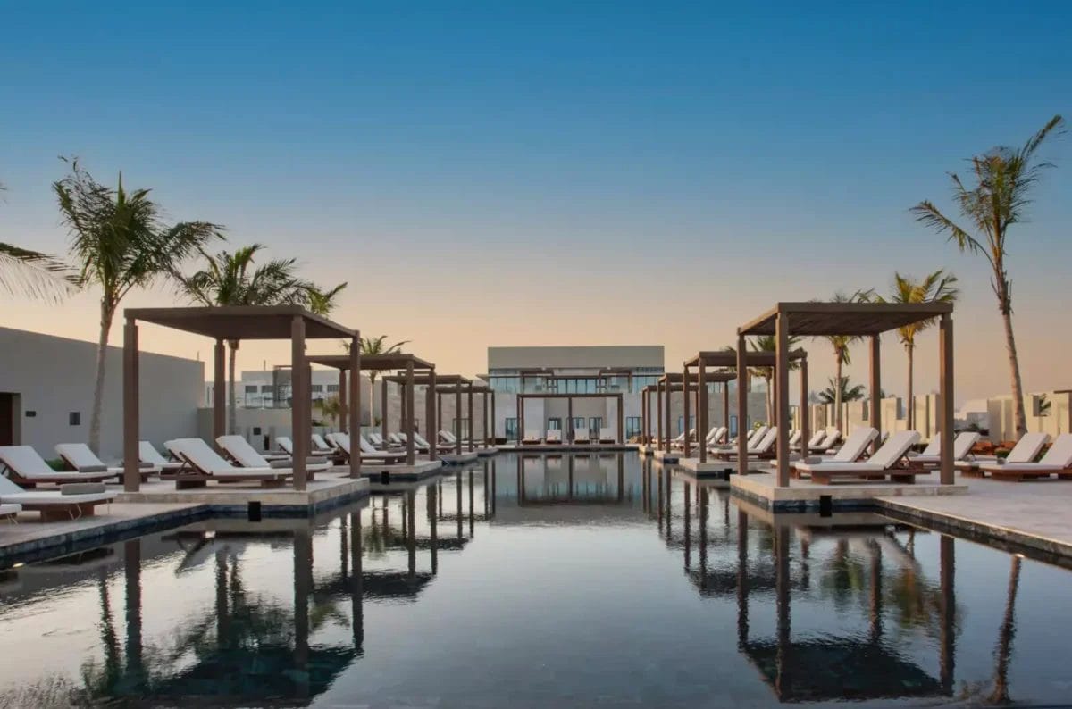 Vivi un'oasi serena ad Alila Hinu Bay, dove una tranquilla piscina è circondata da palme e sedie a sdraio sotto tettoie di legno, il tutto sullo sfondo di un cielo al tramonto mozzafiato. Scopri questa gemma nascosta in Oman Esclusivo per un ritiro indimenticabile.