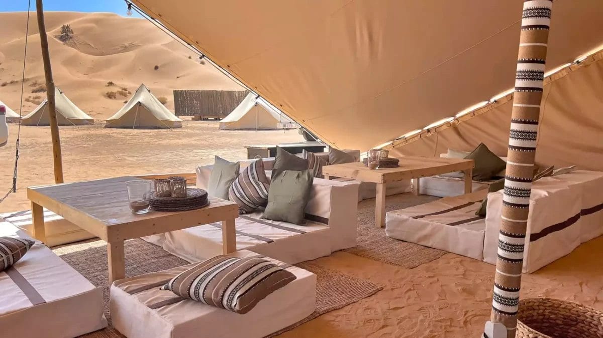 Un'installazione glamping nel deserto con posti a sedere imbottiti, tavoli di legno e cuscini a strisce sotto un grande baldacchino emana un'aria di serenità al Magic Camp. Diverse tende bianche sono visibili sullo sfondo contro il paesaggio sabbioso di Oman Esclusivo.